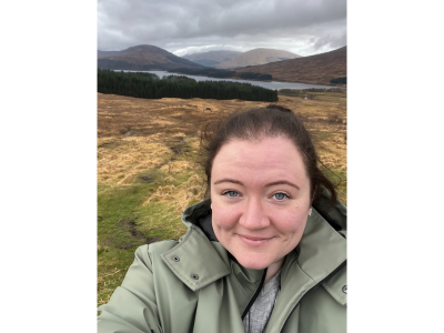 photo of Lauren Louise with countryside and hills in background, white female, long brown hair tied back, wearing pale green jacket