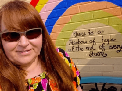 Photograph of Nicola in front of painted rainbow and writing there is a rainbow at the end of every storm. White female with long ginger hair, wearing sunglasses