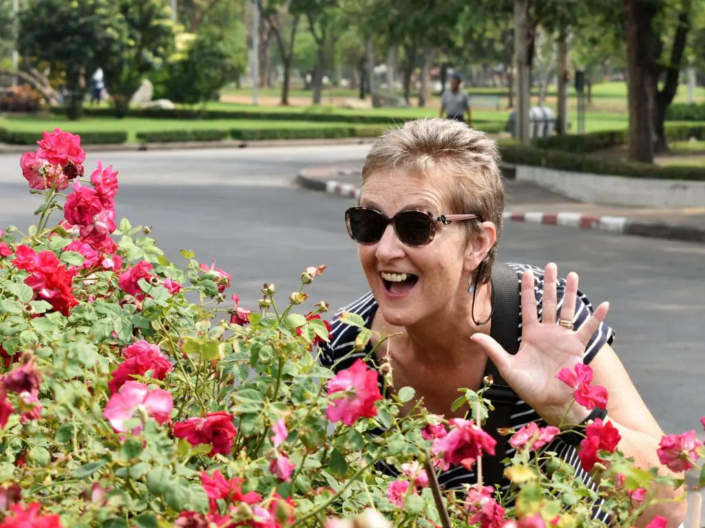 Bev, Peaches supporter, peaking out from behind a rose bush, waving and smiling. White female, short blonde haie, wearing sunglasses and black striped top