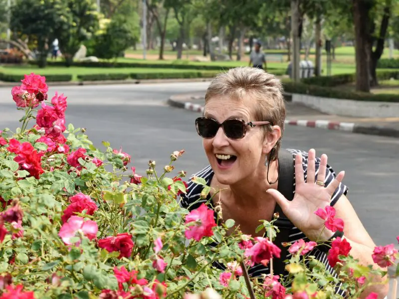 Bev, Peaches supporter, peaking out from behind a rose bush, waving and smiling. White female, short blonde haie, wearing sunglasses and black striped top