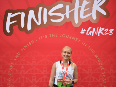 Kate, Peaches fundraiser, standing in front of GNR23 red signage. White female, long blonde hair, holding medal and wearing Peaches running vest