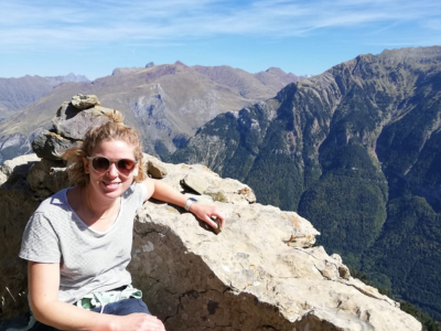 Phillippa, Peaches supporter, crouching with mountain scene in background. White female, blonde hair, wearing sunglasses and pale grey top