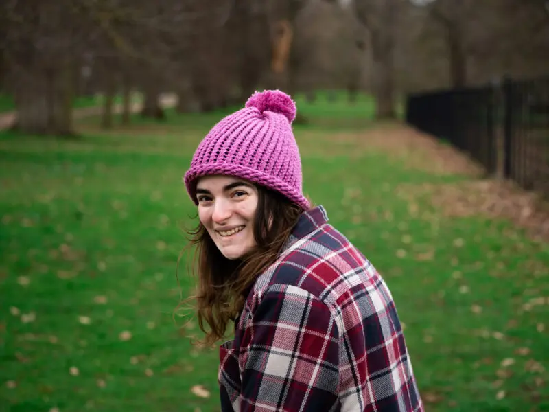 Samantha-Rose, Peaches supporter, sitting in a park with grass and trees in background. White, female wearing pink bobble hat and pink/purple checked shirt